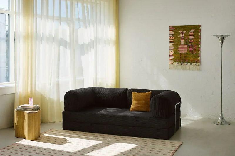 A sleek black corduroy fabric sofa styled in a sunlit Scandinavian interior with natural light, cream curtains, wooden accents, and a woven wall tapestry.